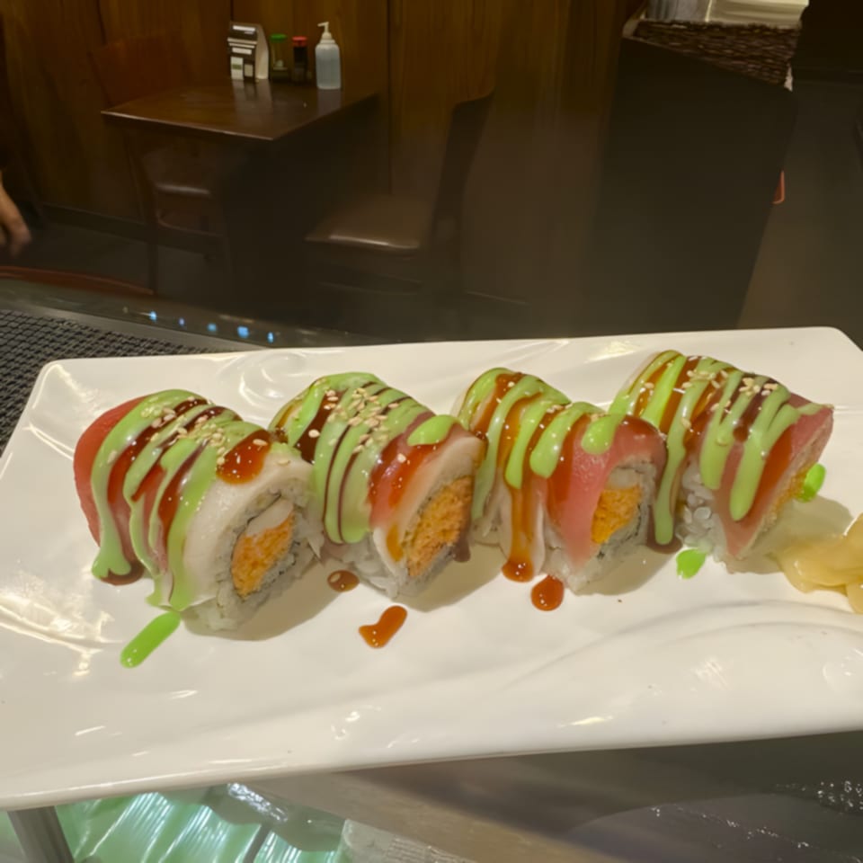 Four Inaka Sushi rolls with green and brown sauce, topped with sesame seeds, on a white plate.