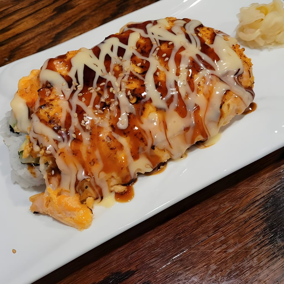 Baked sushi roll with white and brown sauce, sesame seeds, and pickled ginger on a white plate.