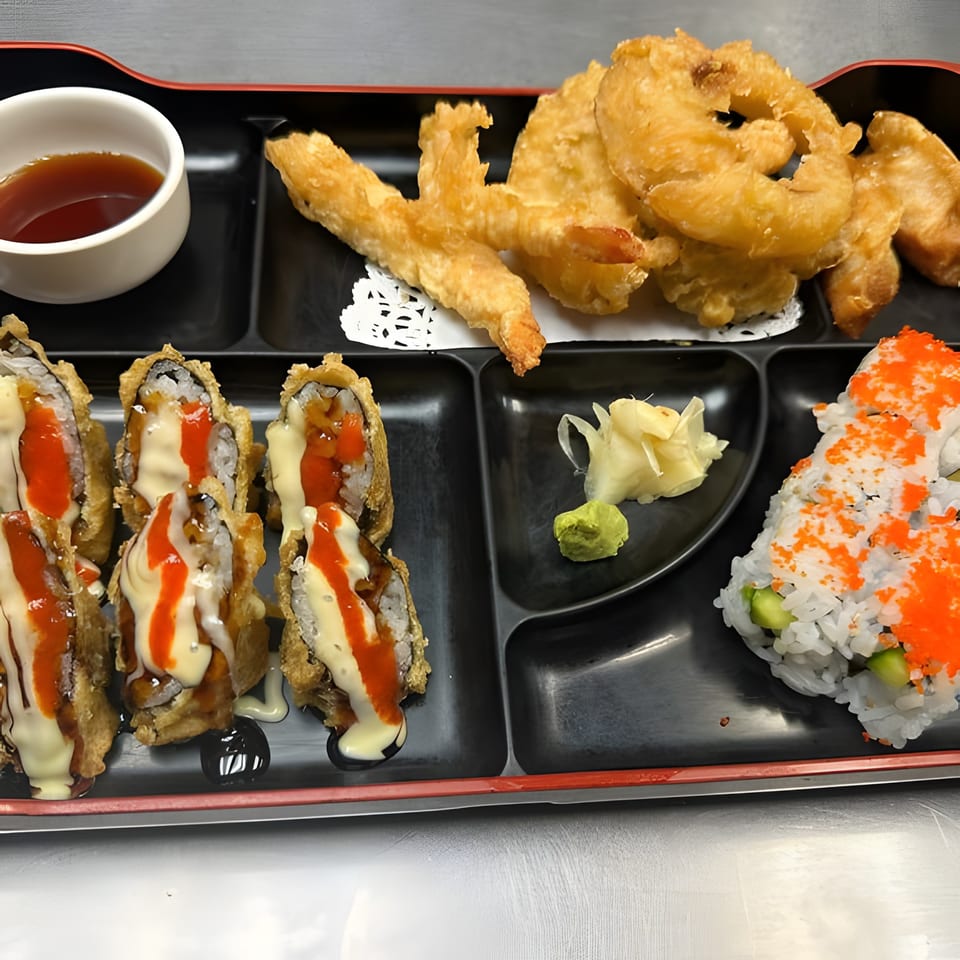 A bento box with tempura shrimp, various sushi rolls, soy sauce, wasabi, and ginger.
