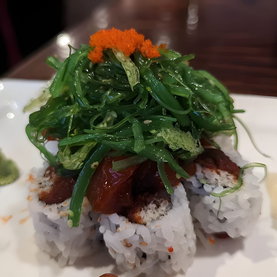 Sushi rolls topped with green seaweed salad, red tuna, and orange tobiko on a white plate.